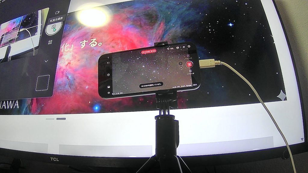 モニターの前に設置された三脚付きのスマートフォン。スマートフォンの画面にはDWARF 3のアプリが表示され、星団のライブ撮影が進行中。背景の大きなモニターには鮮やかなオリオン大星雲のような宇宙の画像が映し出されており、室内からリモートで天体撮影を楽しんでいる様子。